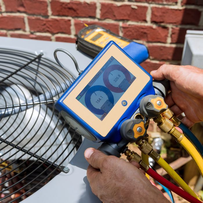 An AC repair tech holding a coolant gauge over an air conditioning unit on the side of a house.